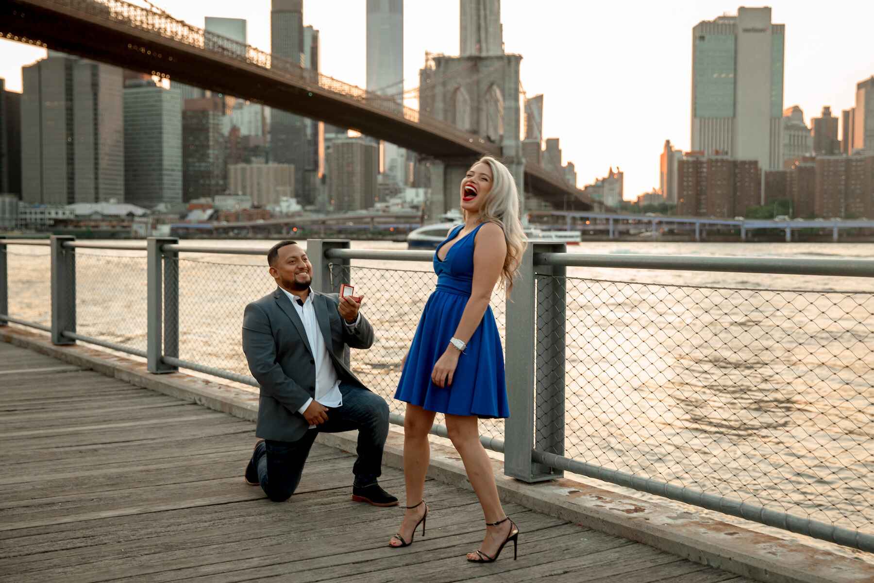 Man proposing to girlfriend wearing blue dress near the Brooklyn Bridge in NYC on a surprise proposal photoshoot