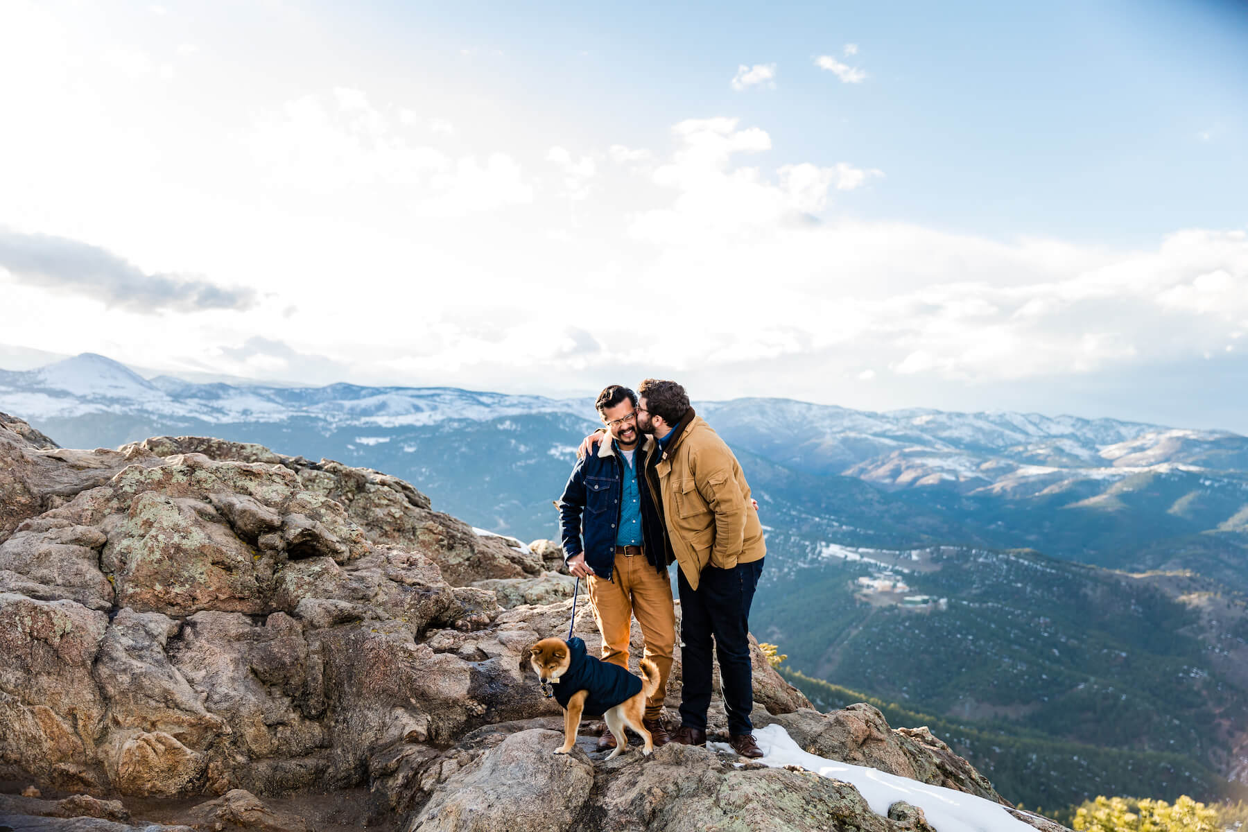 A man kisses his boyfriend on the cheek on the top of a mountain