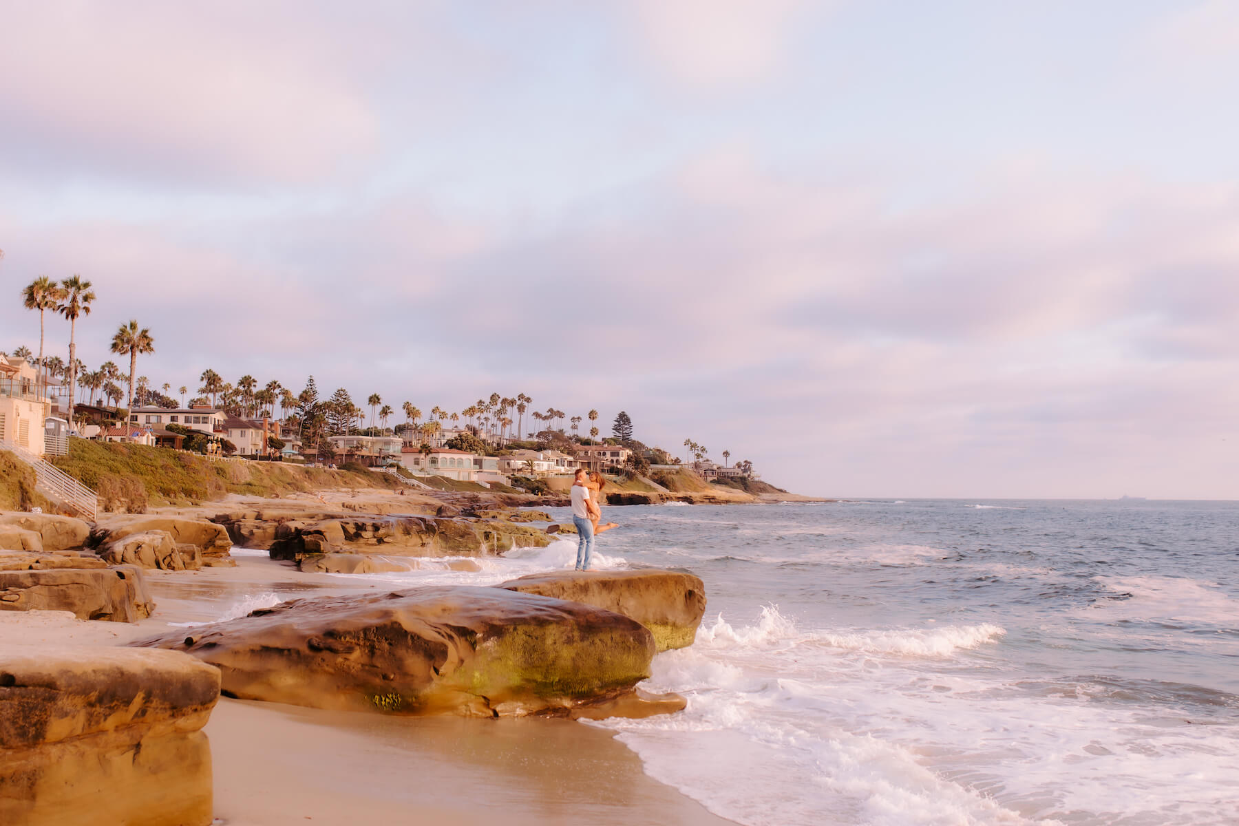 A couple poses dancing at the edge of a large rock appointing into the water in San Diego