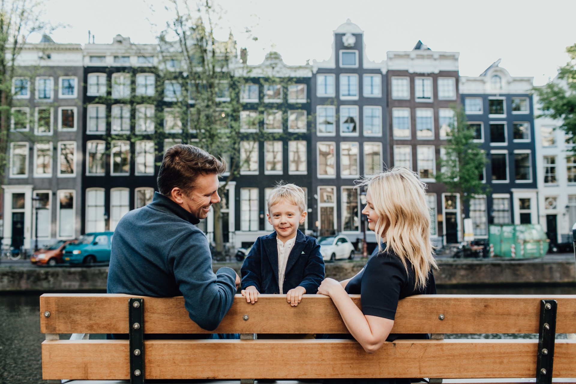Two parents smile at their child on a bench along the canals on their family trip to Amsterdam