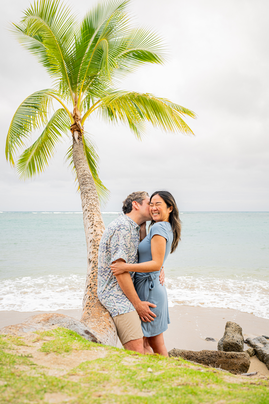 A smiling couple kisses in front of a palm tree on a Honolulu beach on their Flytographer photoshoot.