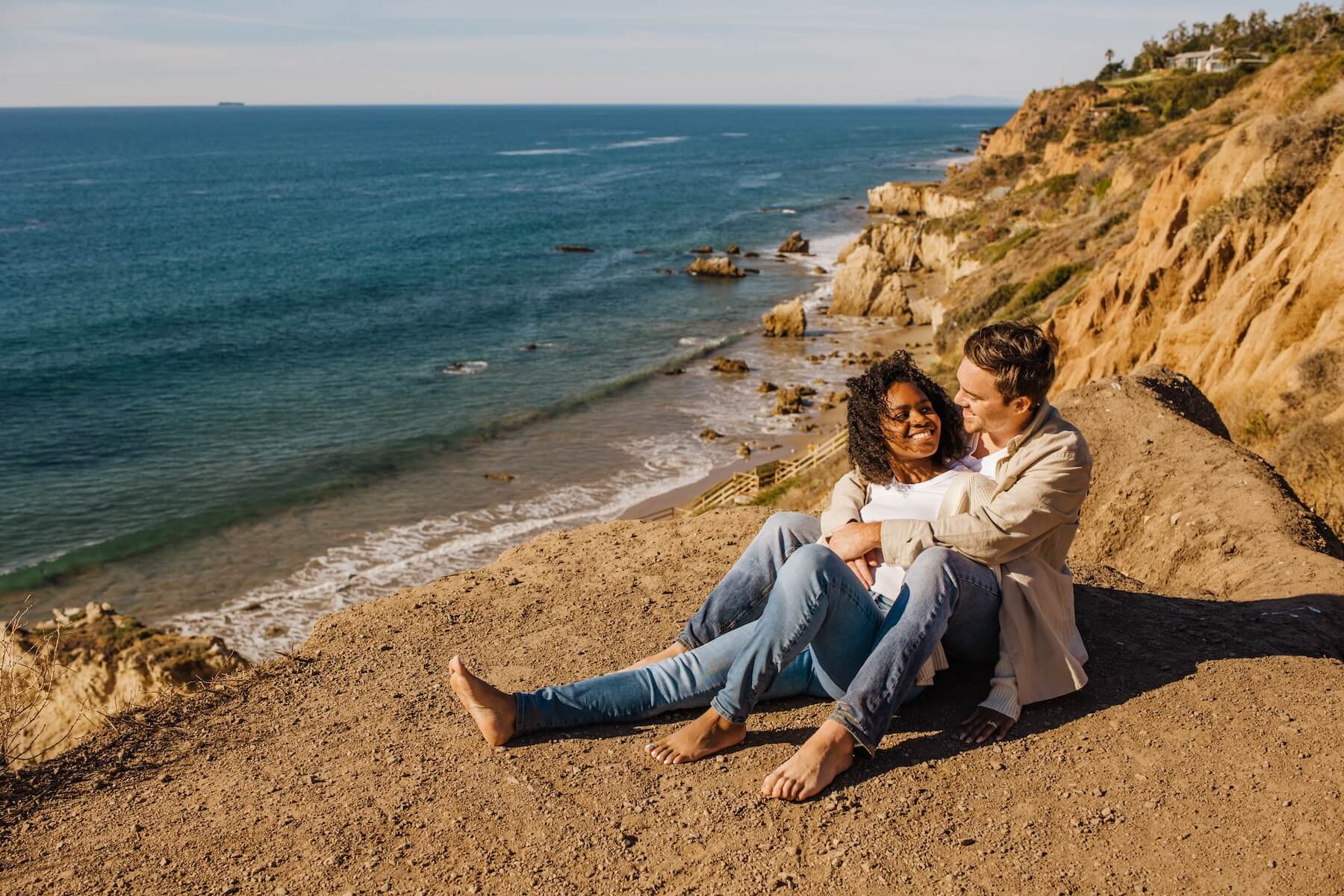 A couple sits together on the edge of a rock looking out at the ocean in Los Angeles California while on an engagement trip 
