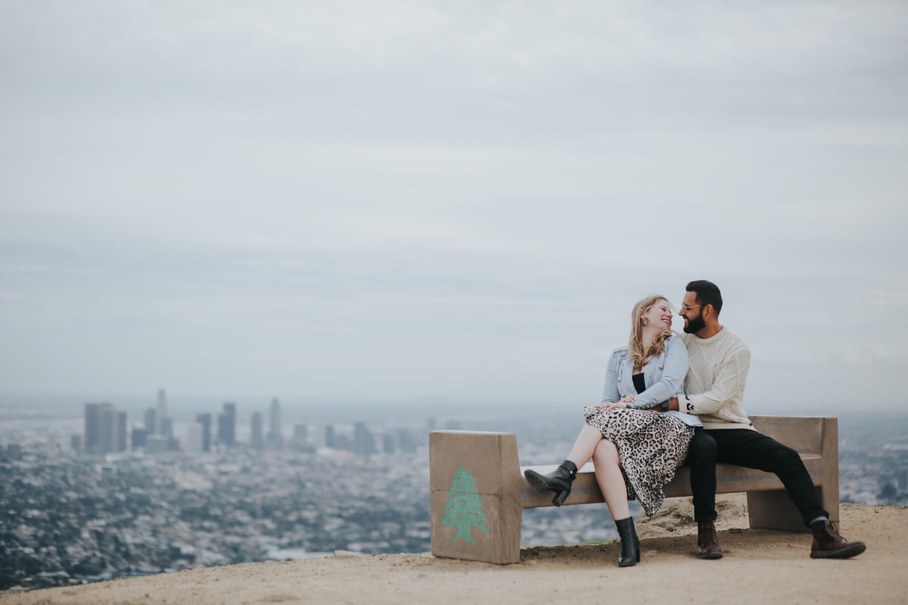A couple sitting on a bench at Hollywood Peak with the Los Angeles skyline in the background