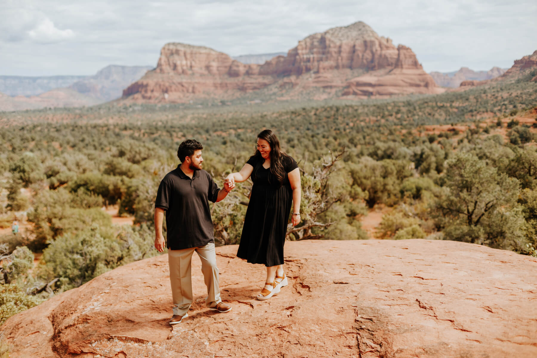 A man and a woman hold hands while walking along a large red rock in Sedona