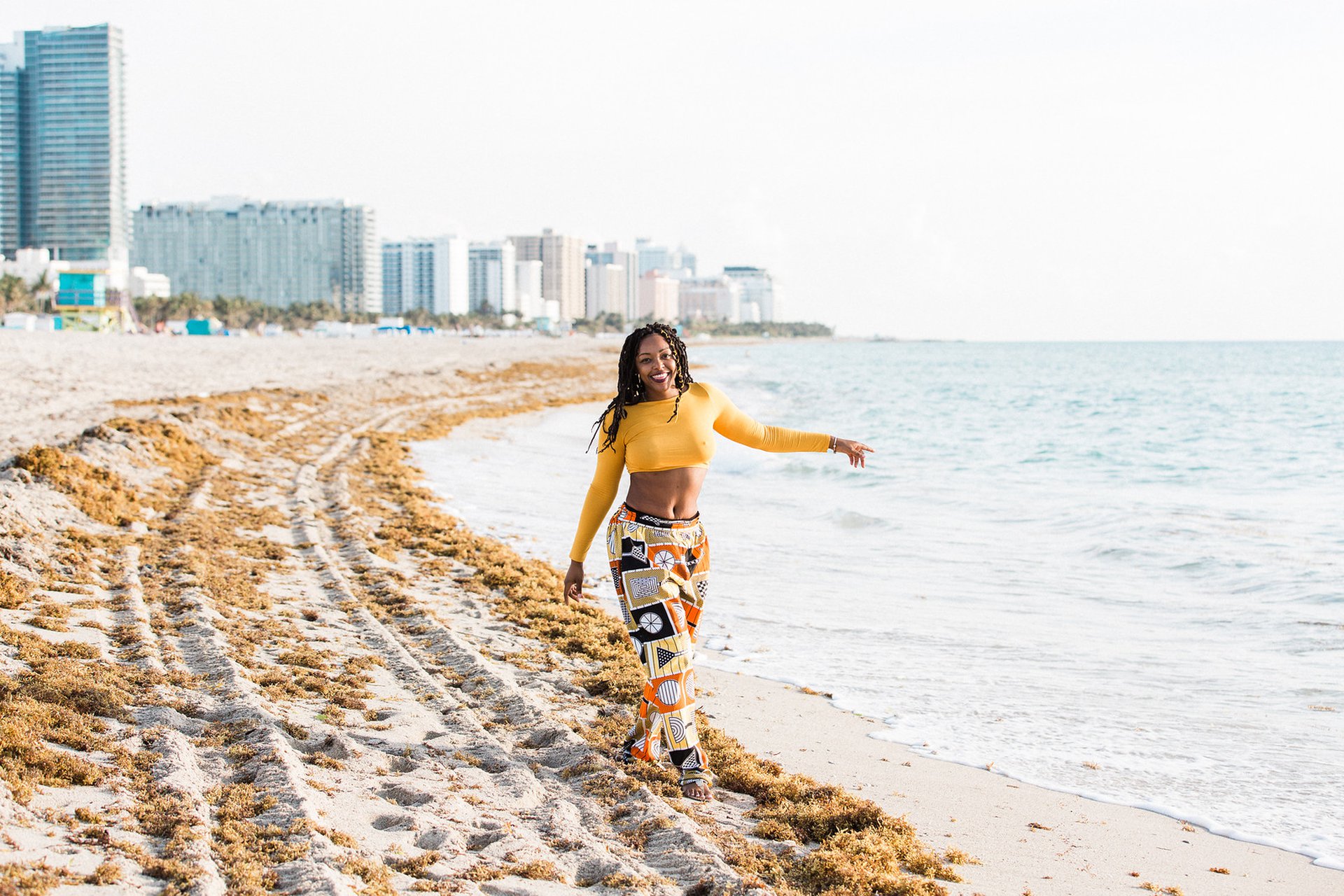 A woman wearing a yellow top and flared pants dancing on the beach in Miami Florida on a photoshoot with Flytographer