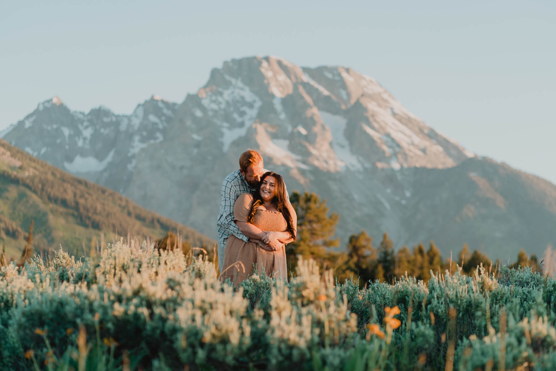 A man hugs a woman from behind in tall grass with mountains in the background