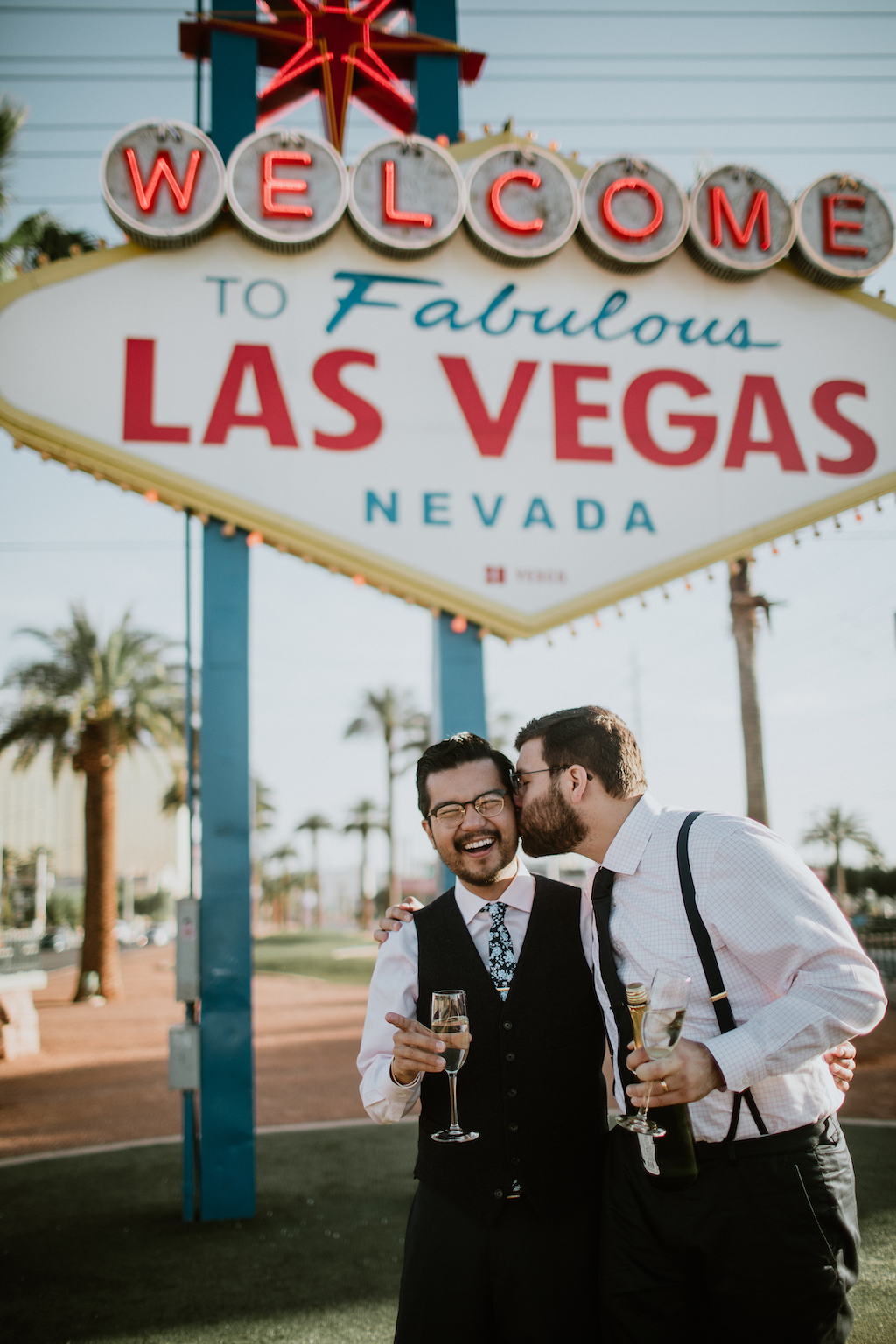A laughing couple kiss with champagne glasses in front of a Las Vegas sign on their Flytographer photoshoot.